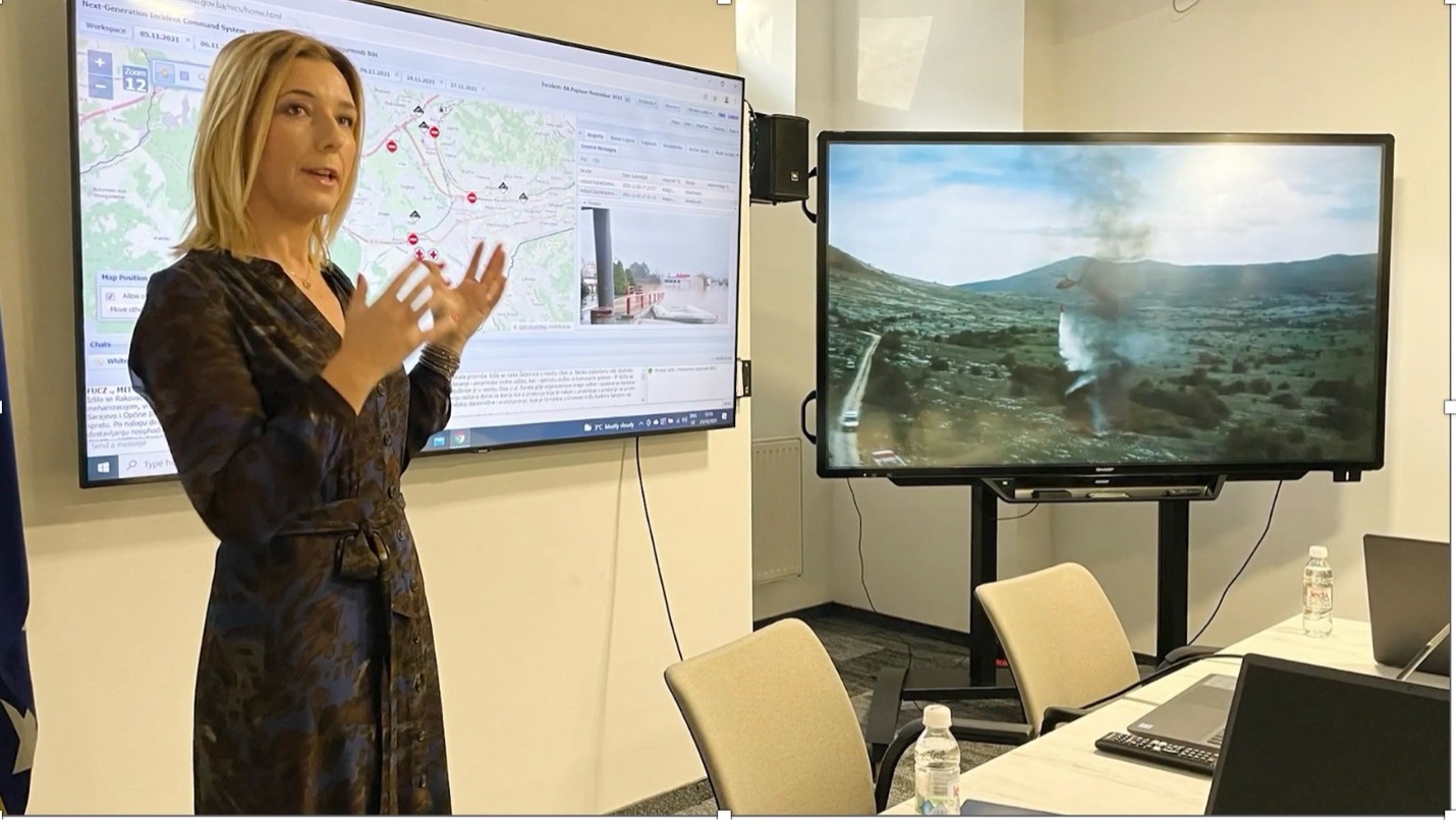 A woman speaks in front of two screens displaying a map and wildfire.
